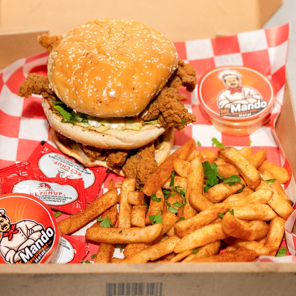 A Mando Halal fried chicken sandwich and seasoned fries in a takeout box with ketchup packets.