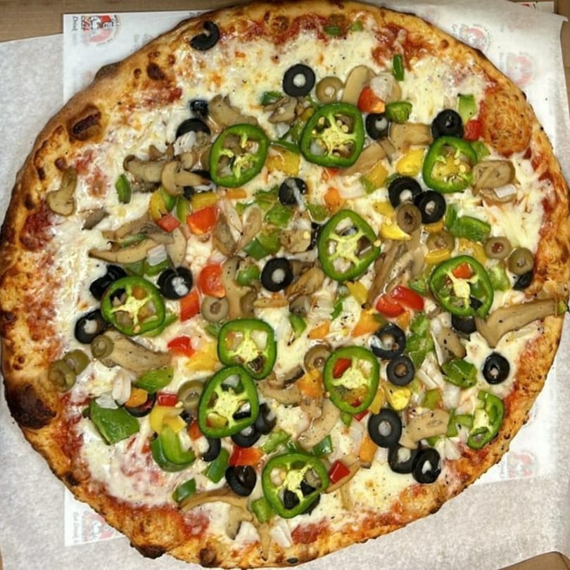 A whole vegetarian pizza with mushrooms, olives, bell peppers, and jalapeños on a white surface.